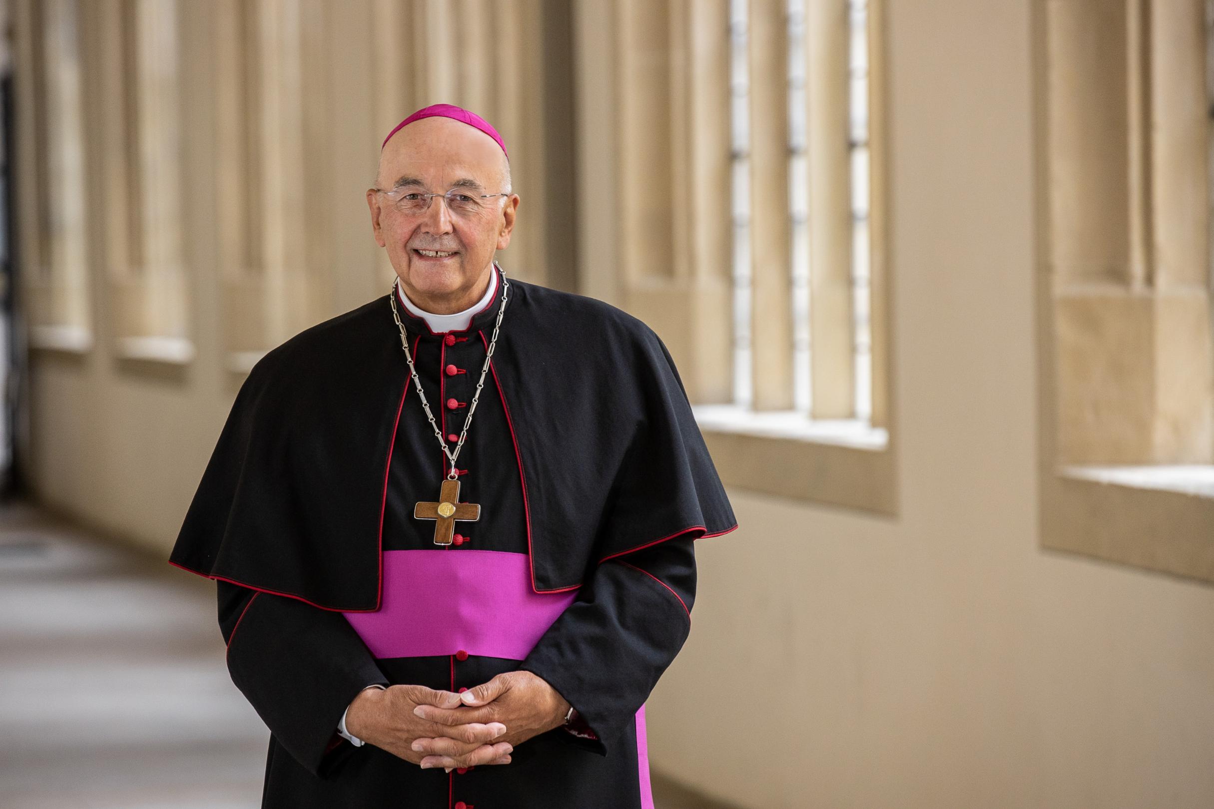 ein Bischof steht in einem Gang welcher beige Mauern hat