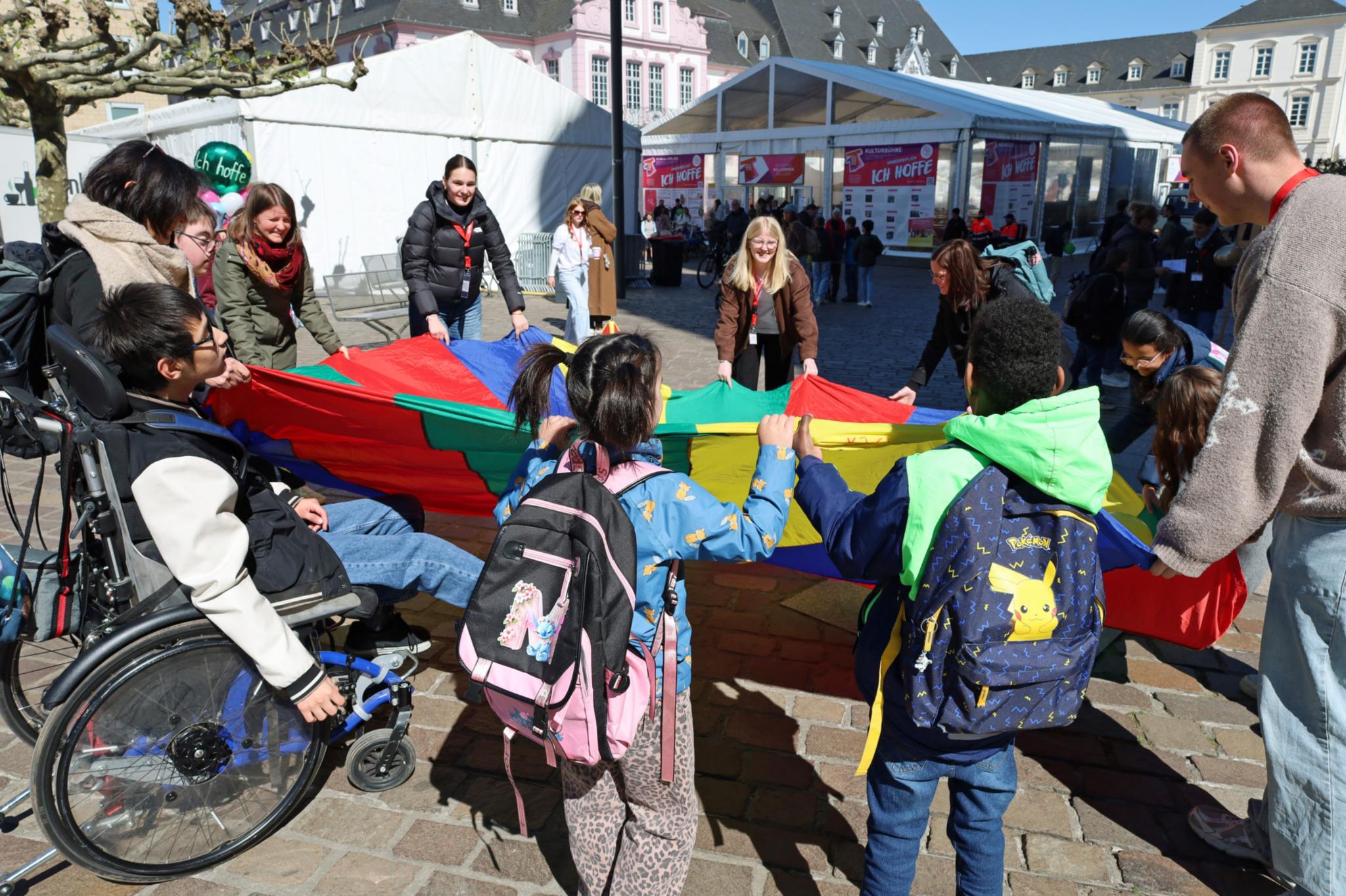 Erwachsene sowie Kinder mit und ohne Beeinträchtigung stehen um ein bundes Tuch herum und ziehen daran.