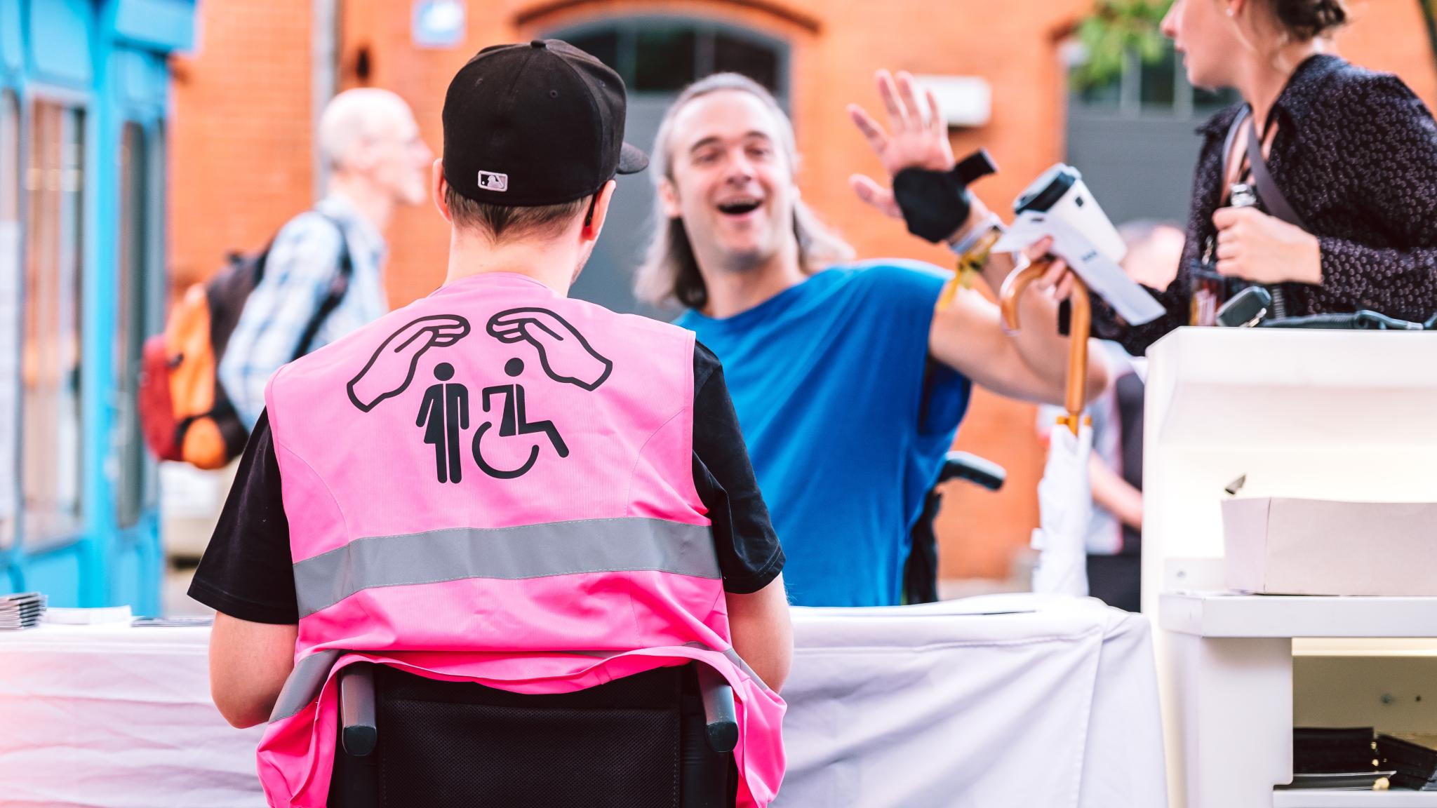 Links im Bild sieht man den Rücken eines Rollstuhlfahrers mit einer pinken Weste. Darauf abgebildet sind zwei Hände, schützend über zwei Symbolen: links das für All gender welcome und rechts das für Rollstuhlfahrer*innen. Im Hintergrund sitzt ein junger Mann in blauem T-Shirt, ebenfalls Rollstuhlfahrer, der in die Kamera winkt und lächelt.