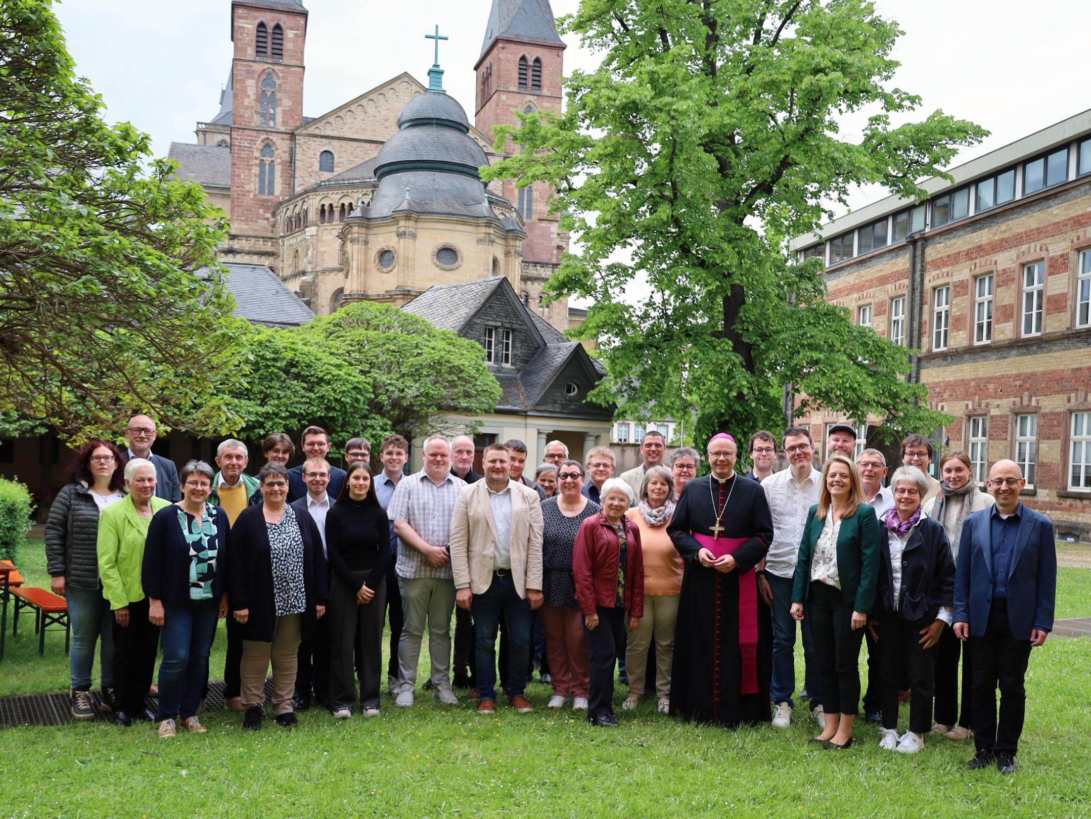 Eine Gruppe von Menschen steht zusammen mit Bischof Ackermann auf einer Wiese. Im Hintergrund ist der Dom